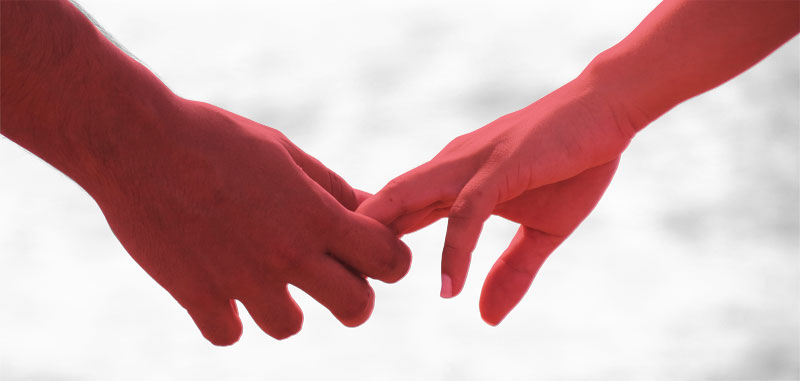Primer plano de dos manos acercándose con las yemas de los dedos casi tocándose, en tonos rojos y rosados con fondo suave y difuso, evocando cercanía y apoyo